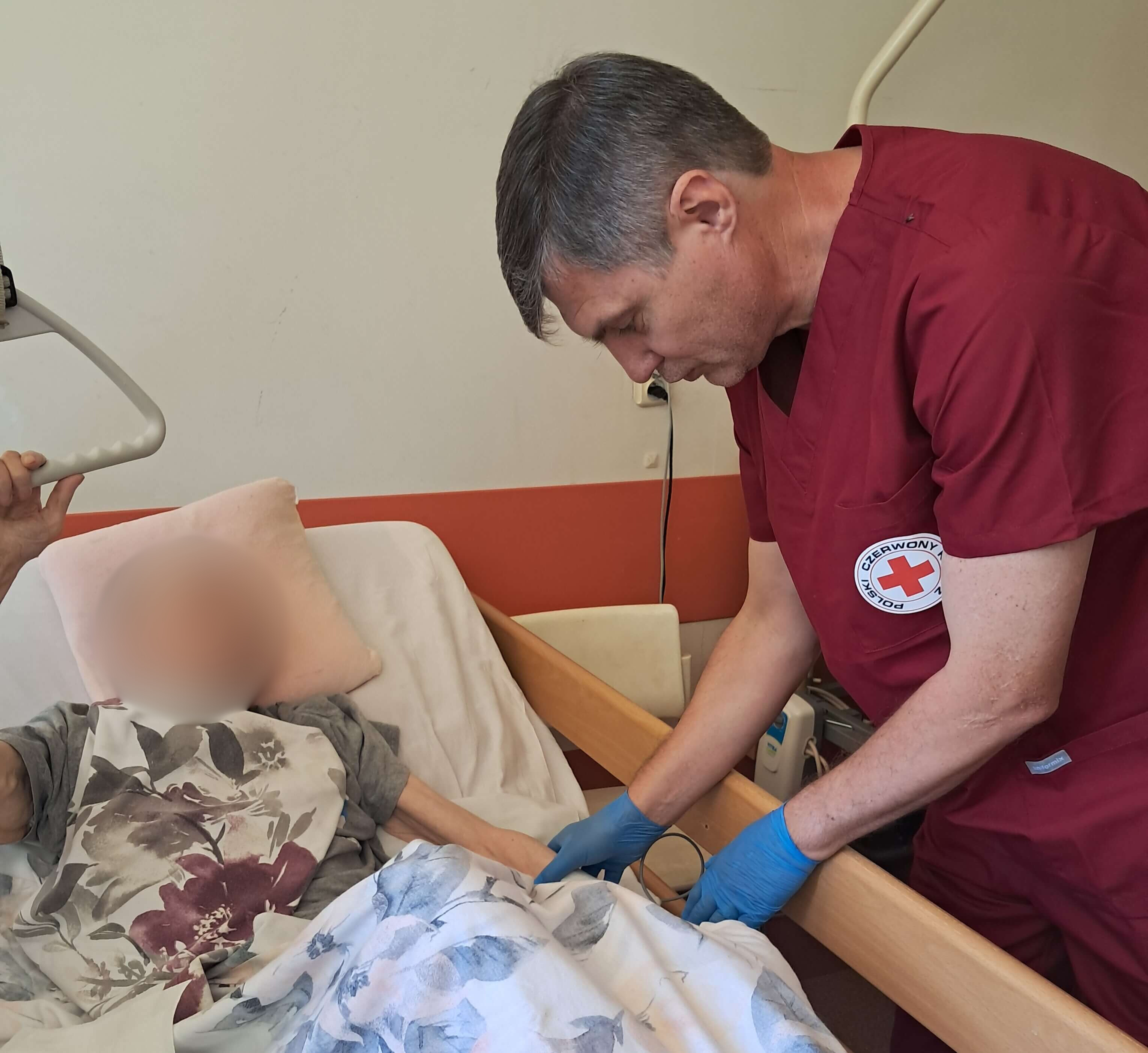 Opiekun medyczny w bordowym uniformie z logo Polskiego Czerwonego Krzyża pomaga starszej osobie leżącej w łóżku szpitalnym.