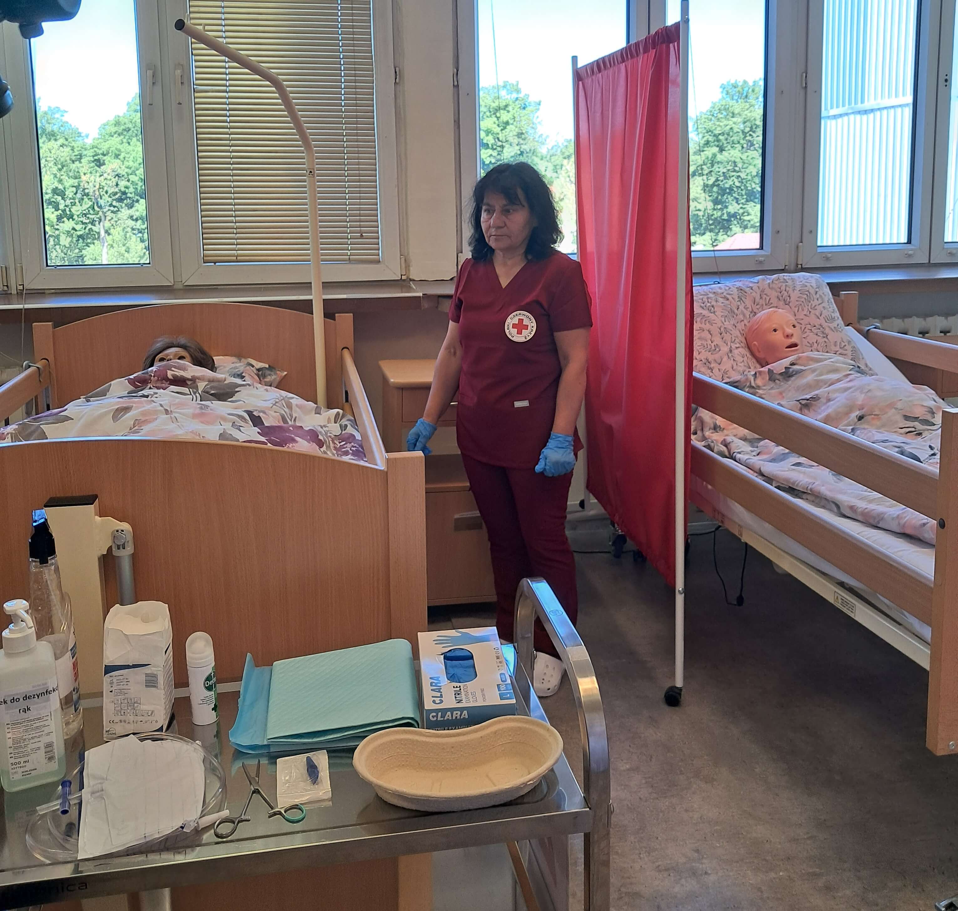 Kobieta w bordowym uniformie z logo Polskiego Czerwonego Krzyża stoi przy łóżkach w sali opieki.