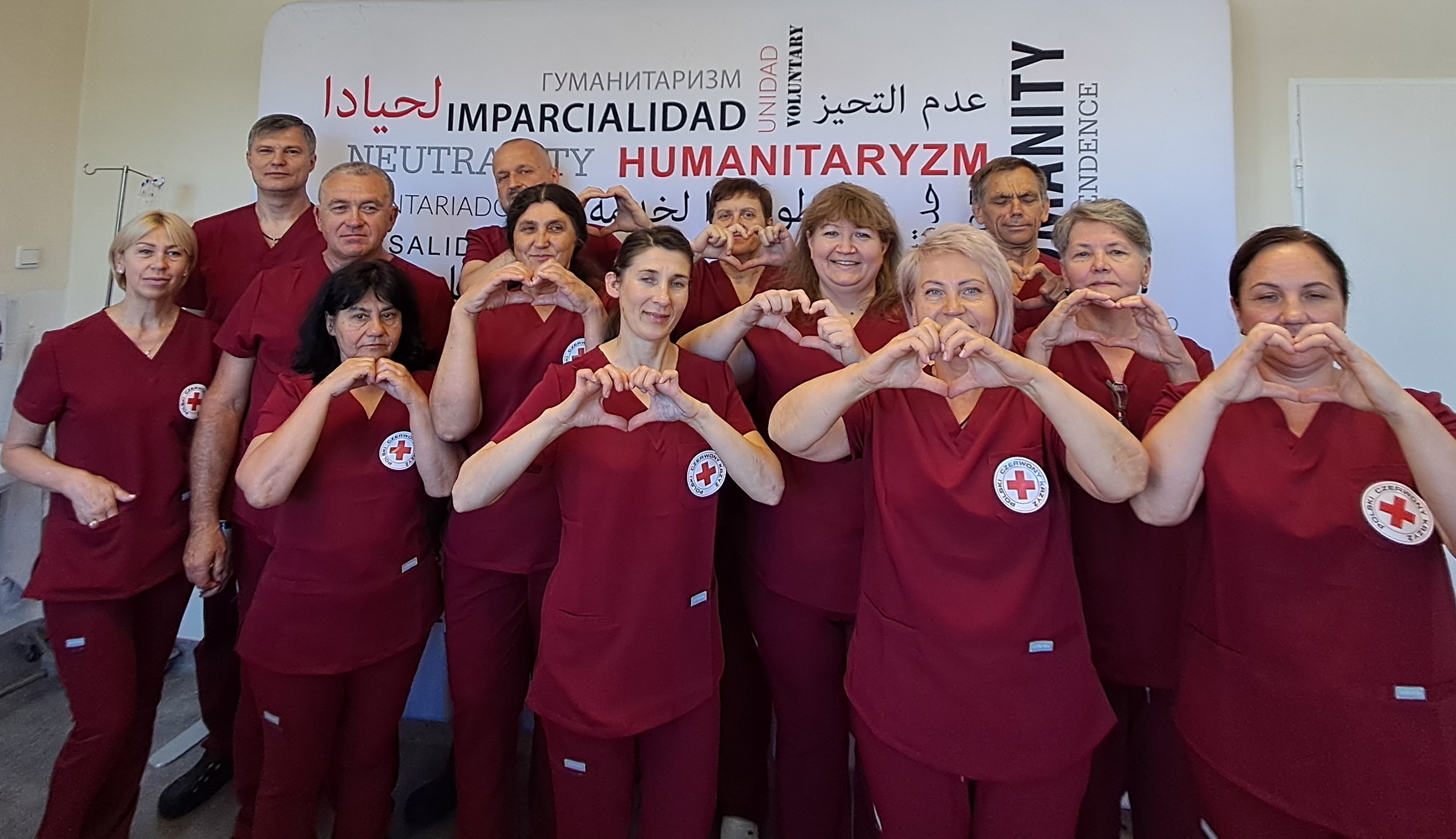 Osoby ubrane w szpitalne uniformy z uśmiechami na twarz unoszą dłonie, których zetknięte palce formują kształy serc.